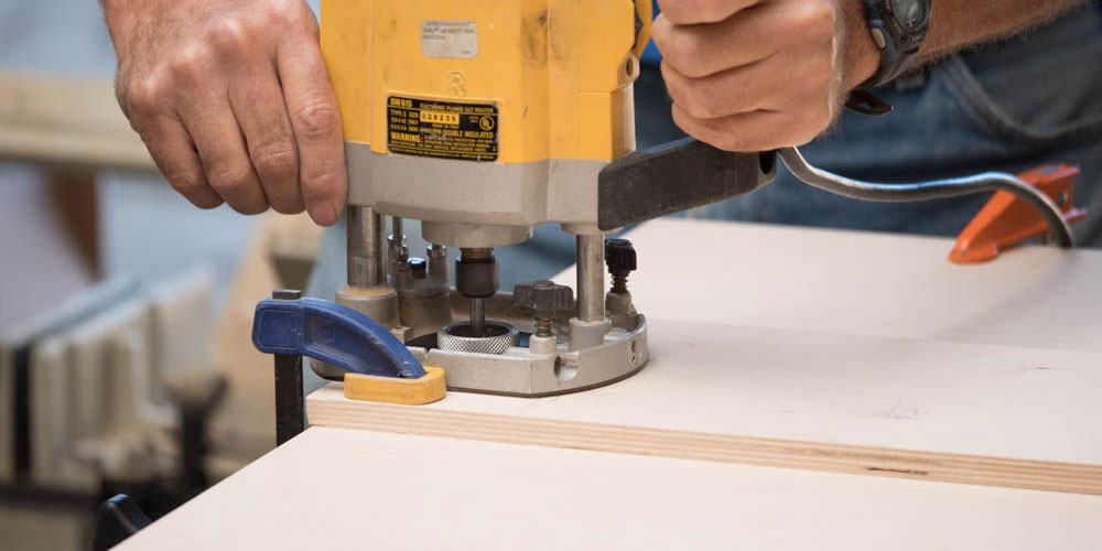 A pair of hands using an electric router to machine wood parts.